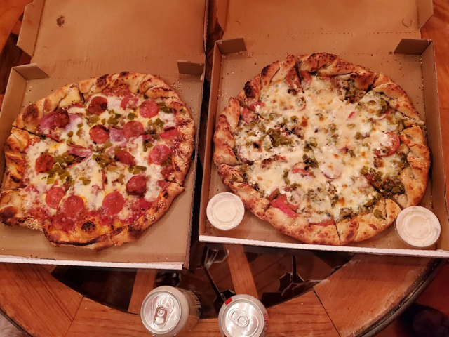 Two takeout pizzas side by side, one pepperoni and one loaded with green chile