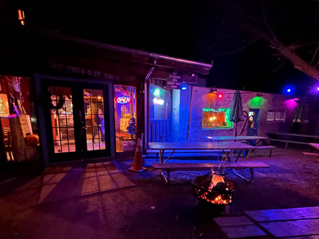 Neon-lit entrance at night with a fire pit glowing beside the picnic tables