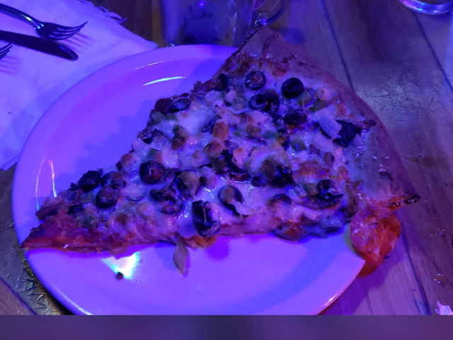 Slice of black olive and green chile pizza on a plate under purple neon light